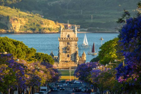 Belem Tower, Jacaranda Blooming Purple Blue Trees and Sailboats on Sunny Evening. Golden Hour. Lisbon, Portugal.