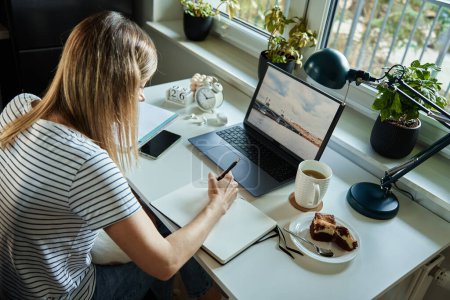 Foto de Freelancer trabajando en casa. Mujer usando portátil y escribiendo en bloc de notas sentado en la mesa en la oficina en casa - Imagen libre de derechos
