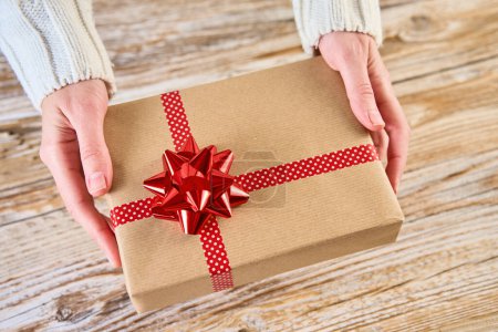 Photo for Woman holds gift box wrapped in craft paper with festive ribbon agains wooden background. Female hands holding present for christmas holidays, above view - Royalty Free Image