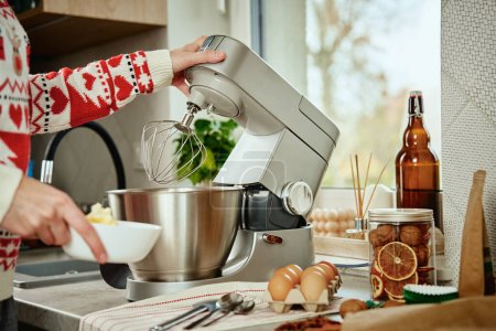 Woman in festive christmas sweater preparing dough for cookies at home kitchen. Female hands use electric mixer. Modern kitchen household appliances