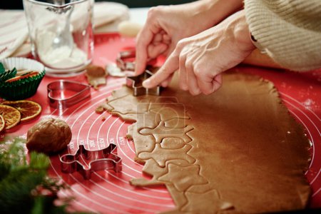 Foto de Mujer preparando galletas de jengibre en la cocina con decoraciones navideñas. Manos femeninas cortando masa de jengibre con cortador para hacer galletas para las vacaciones de invierno - Imagen libre de derechos