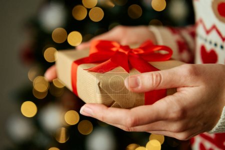 Téléchargez les photos : Femme en pull chaud tenant boîte cadeau enveloppant dans du papier artisanal avec ruban festif. Présent pour les vacances d'hiver. Noël et bonne année célébrant - en image libre de droit