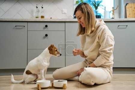 Foto de Mujer poniendo tazón de comida con pienso para su perro en el suelo en la cocina, propietaria pasando tiempo junto con su mascota en casa, alimentación de animales y cuidado de mascotas - Imagen libre de derechos