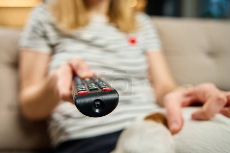 Woman holds TV remote control. Female is sitting on sofa in living room and switching channels on the TV. Concept of entertainment and lazy weekends