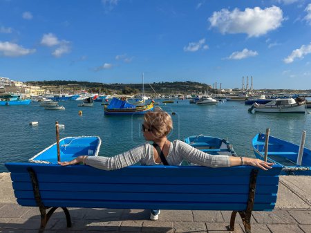 Woman sitting on bench and looking at boats in Marsaxlokk harbor, Malta