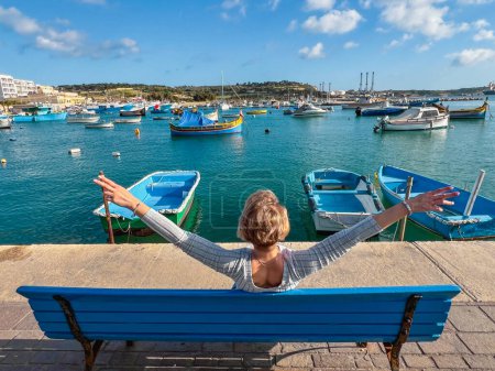 Woman sitting on bench and looking at boats in Marsaxlokk harbor, Malta