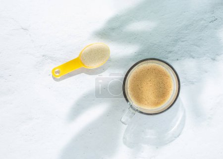 Téléchargez les photos : Sunlit cup of coffee with spoonful of collagen on isolated pastel white background with shadows of monstera leaf. Aesthetic concept of dietary supplements for health and beauty. Minimal flat lay. - en image libre de droit
