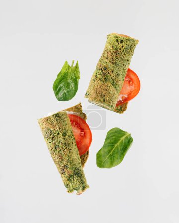 Spinatblätter und gerollte grüne Tortilla oder Pfannkuchen halbiert, mit Tomatenscheiben, die über isoliertem pastellweißem Hintergrund schweben. Minimales Konzept ketogener Kost oder vegetarisches Low-Carb-Frühstück.