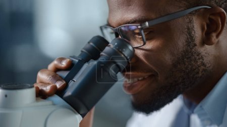Foto de Macro Primer plano de un guapo científico negro con gafas y mirando al microscopio. Microbiólogo trabajando en muestras moleculares en laboratorio moderno con equipo tecnológico. - Imagen libre de derechos