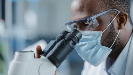 Photo for Close Up Shot of a Handsome Black Male Scientist Wearing Face Mask and Looking into the Microscope. Microbiologist Working on Molecule Samples in Modern Laboratory with Technological Equipment. - Royalty Free Image