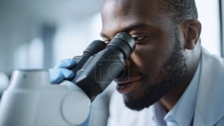 Téléchargez les photos : Macro Close Up Shot of a Handsome Excited Black Male Scientist Looking into the Microscope. Microbiologiste travaillant sur des échantillons de molécules en laboratoire moderne avec équipement technologique. - en image libre de droit