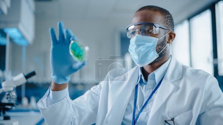 Téléchargez les photos : Homme scientifique noir portant masque facial et lunettes regardant Petri plat avec des produits chimiques d'échantillon génétiquement modifiés. Microbiologiste travaillant dans un laboratoire moderne avec des équipements technologiques. - en image libre de droit