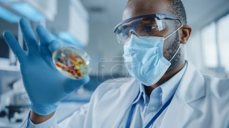Foto de Científico masculino negro que usa mascarilla facial y gafas mirando el plato Petri con productos químicos genéticamente modificados de la muestra. Microbiólogo Trabajando en Laboratorio Moderno con Equipo Tecnológico. - Imagen libre de derechos