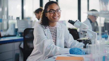 Photo for Medical Science Laboratory: Beautiful Black Scientist is Using Microscope, Looking at Camera and Smiling Charmingly. Young Biotechnology Science Specialist, Using Technologically Advanced Equipment. - Royalty Free Image