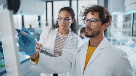 Photo for Medical Science Laboratory: Handsome Latin Male Scientist Writes Detailed Project Data Analysis on the Board, His Black Female Colleague Talks. Young Scientists Solving Problems. - Royalty Free Image