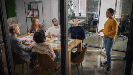 Photo for Project Manager Makes a Presentation for a Young Diverse Creative Team in Meeting Room in an Agency. Colleagues Sit Behind Conference Table and Discuss Business Development, User Interface and Design. - Royalty Free Image