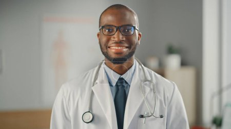 Téléchargez les photos : Fermer Portrait d'un médecin de famille afro-américain heureux portant des lunettes dans une clinique de santé. Réussi médecin noir en blouse blanche regarde la caméra et sourit dans le bureau de l'hôpital. - en image libre de droit
