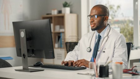 Photo for Experienced African American Male Doctor Wearing White Coat Working on Personal Computer at His Office. Medical Health Care Professional Working with Test Results, Patient Treatment Planning. - Royalty Free Image