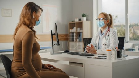 Foto de La doctora de familia está hablando con una joven embarazada durante la consulta en una clínica de salud. Ambos usan máscaras faciales. Médico en bata de laboratorio sentado detrás de un escritorio de computadora en la oficina del hospital. - Imagen libre de derechos
