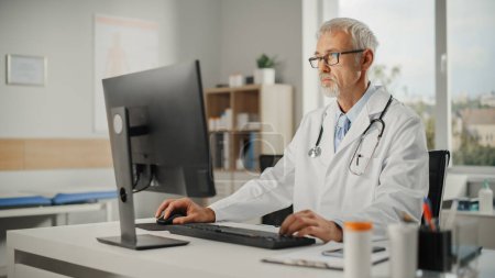 Photo for Experienced Middle Aged Male Doctor Wearing White Coat Working on Personal Computer at His Office. Senior Medical Health Care Professional Working with Test Results, Patient Treatment Planning. - Royalty Free Image