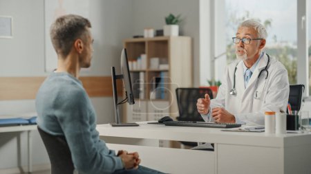Photo for Middle Aged Family Doctor is Talking with Young Male Patient During Consultation in a Health Clinic. Senior Physician in Lab Coat Sitting Behind a Computer Desk in Hospital Office. - Royalty Free Image