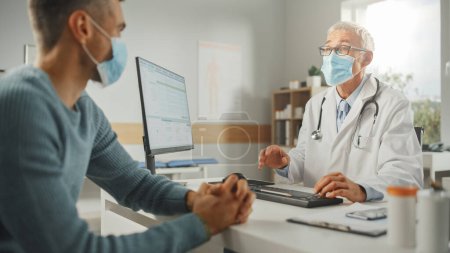 Photo for Middle Aged Family Doctor is Talking with Young Male Patient During Consultation in a Health Clinic. Both Wear Face Masks. Physician in Lab Coat Sitting Behind a Computer Desk in Hospital Office. - Royalty Free Image