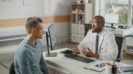 Photo for African American Family Doctor is Talking with Young Male Patient During Consultation in a Health Clinic. Black Physician in Lab Coat and Glasses Sitting Behind a Computer Desk in Hospital Office. - Royalty Free Image