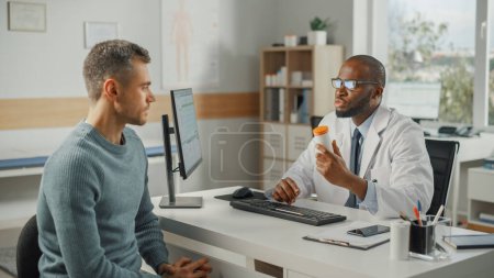 Photo for African American Family Doctor is Talking with Young Male Patient During Consultation in a Health Clinic. Black Physician in Lab Coat and Glasses Sitting Behind a Computer Desk in Hospital Office. - Royalty Free Image