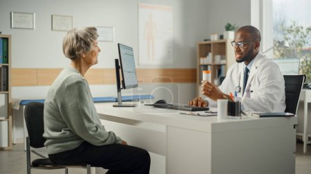 Photo for African American Family Doctor is Prescribing Medication to Senior Female Patient and Speaking with Her During Consultation in a Health Clinic. Physician Sitting Behind a Desk in Hospital Office. - Royalty Free Image