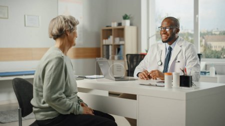 Téléchargez les photos : Le médecin de famille afro-américain prescrit des médicaments à une patiente âgée et lui parle pendant une consultation dans une clinique de santé. Médecin assis derrière un bureau dans le bureau de l'hôpital. - en image libre de droit