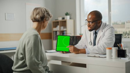 Téléchargez les photos : Médecin de famille afro-américain montrant Tablet Computer avec écran vert à la patiente âgée pendant la consultation dans une clinique de santé. Médecin assis derrière un bureau dans le bureau de l'hôpital. - en image libre de droit