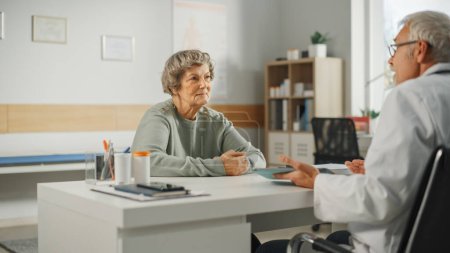 Photo for Middle Aged Family Doctor is Talking with Senior Female Patient During Consultation in a Health Clinic. Experienced Caucasian Physician in Lab Coat Sitting Behind a Computer Desk in Hospital Office. - Royalty Free Image