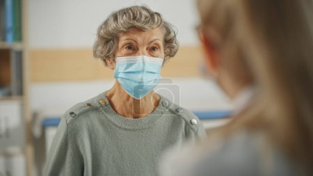 Téléchargez les photos : Une femme médecin de famille s'adresse à une femme âgée dans une clinique de santé. Les deux portent un masque facial. Médecin en blouse de laboratoire prescrivant des médicaments aux patients âgés au bureau de l'hôpital. - en image libre de droit