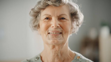 Téléchargez les photos : Authentique Gros plan Portrait d'une retraitée âgée dans une pièce lumineuse à la maison. Belle vieille femelle avec des poses de cheveux gris pour l'appareil photo et sourit doucement. Bonne personne âgée pleine de santé. - en image libre de droit