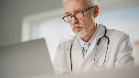 Photo for Experienced Middle Aged Male Doctor Wearing White Coat Working on Laptop Computer at His Office. Senior Medical Health Care Professional Working with Test Results, Patient Treatment Planning. - Royalty Free Image