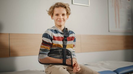 Photo for Portrait of a Healthy Young Handsome Teenage Boy Sitting on a Bench with His Hand in an Arm Brace in a Health Clinic. Child in Colorful Jumper is Happy and Smiling in Hospital Office. - Royalty Free Image