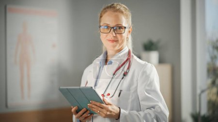 Téléchargez les photos : Portrait d'une femme médecin expérimentée portant un manteau blanc travaillant sur tablette à son bureau. Un professionnel de la santé ou une infirmière en chef regarde la caméra et sourit doucement. - en image libre de droit