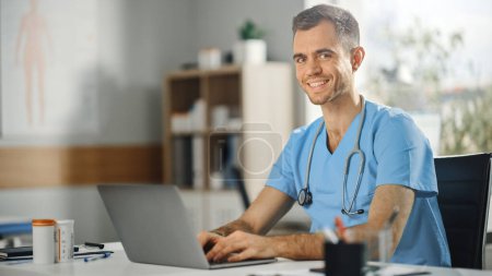 Téléchargez les photos : Infirmière de sexe masculin portant l'uniforme bleu travaillant sur ordinateur portable au bureau du médecin et souriant à la caméra. Professionnel de la santé travaillant sur la lutte contre les stéréotypes à la diversité des sexes dans la carrière infirmière. - en image libre de droit