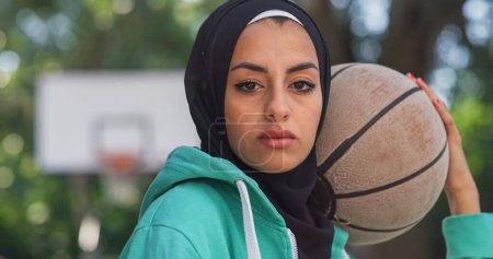 Téléchargez les photos : Gros plan de la belle jeune femme musulmane tenant un ballon de basket tout en regardant la caméra. Athlète féminine dans un hijab défiant les stéréotypes et suivant son rêve de devenir professionnelle - en image libre de droit