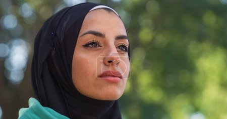 Téléchargez les photos : Portrait de belle jeune femme arabe regardant dans l'horizon avec un fond de verdure. Athelette musulmane avec hijab profitant de l'air frais dans le parc et prête à voir des amis - en image libre de droit