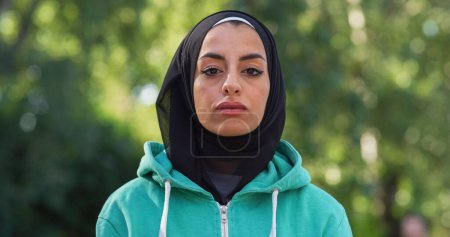 Téléchargez les photos : Portrait de belle jeune femme arabe avec un fond de verdure regardant la caméra. Athelette musulmane heureuse avec hijab profitant de l'air frais dans le parc pendant son entraînement tôt le matin - en image libre de droit