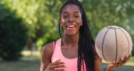 Téléchargez les photos : Portrait d'une jeune femme noire forte lançant un ballon de basket tout en regardant la caméra dans une cour extérieure. Athlète féminine souriante défiant les stéréotypes et suivant son rêve de devenir professionnelle - en image libre de droit
