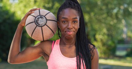 Téléchargez les photos : Portrait d'une jeune femme noire forte tenant un ballon de basket tout en regardant la caméra dans une cour extérieure. Athlète féminine souriante défiant les stéréotypes et suivant son rêve de devenir professionnelle - en image libre de droit