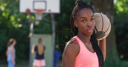 Téléchargez les photos : Portrait d'une belle jeune femme noire tenant un ballon de basket tout en regardant la caméra dans une cour extérieure. Athlète féminine défiant les stéréotypes et suivant son rêve de devenir professionnelle - en image libre de droit