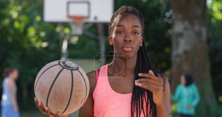 Téléchargez les photos : Portrait d'une belle jeune femme noire lançant un ballon de basket tout en regardant la caméra dans une cour extérieure. Athlète féminine défiant les stéréotypes et suivant son rêve de devenir professionnelle - en image libre de droit