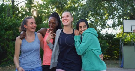 Foto de Retrato de amigas multiétnicas en ropa deportiva Abrazando y riendo. Grupo de mujeres jóvenes celebrando una victoria en la cancha de baloncesto al aire libre juntas, contando chistes y disfrutando de la juventud - Imagen libre de derechos