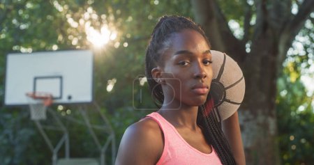 Téléchargez les photos : Portrait d'une belle jeune femme noire tenant un ballon de basket tout en regardant la caméra dans une cour extérieure. Athlète féminine défiant les stéréotypes et suivant son rêve de devenir professionnelle - en image libre de droit