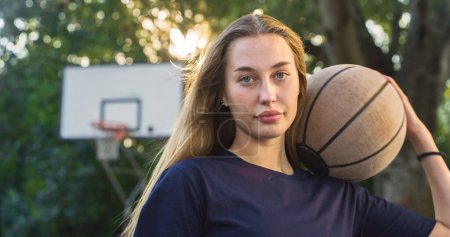 Téléchargez les photos : Portrait d'une belle jeune femme blonde tenant un ballon de basket tout en regardant la caméra dans un tribunal extérieur. Athlète féminine défiant les stéréotypes et suivant son rêve de devenir professionnelle - en image libre de droit