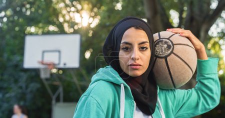 Téléchargez les photos : Gros plan de la belle jeune femme musulmane tenant un ballon de basket tout en regardant la caméra. Athlète féminine dans un hijab défiant les stéréotypes et suivant son rêve de devenir professionnelle - en image libre de droit