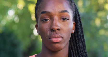 Téléchargez les photos : Gros plan de belle jeune femme noire regardant la caméra avec une expression neutre avec un fond de verdure. Adolescente heureuse profitant de l'air frais dans le parc et prenant soin de son bien-être - en image libre de droit